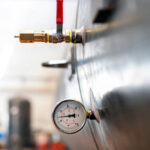 tank-Pressure-test-petromechzw Close-up of a pressure gauge and valve attached to the side of a large metal tank in an industrial setting, with blurred equipment and PLC controls visible in the background.