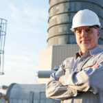 A serious, mature man, in his 50s, working at a power station, standing outdoors with his arms folded, wearing a gray uniform, white hardhat and safety glasses.  We see the manufacturing facility in the background.