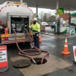 A worker in safety gear transfers fuel from a tanker truck into an underground tank at a gas station, surrounded by orange cones and signs reading FLAMMABLE, FUEL TRANSFER IN PROGRESS, and NO ENTRY.