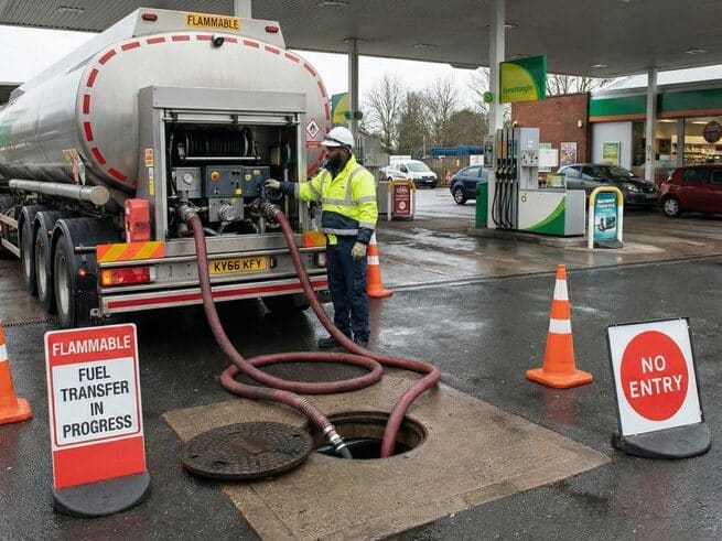 A worker in safety gear transfers fuel from a tanker truck into an underground tank at a gas station, surrounded by orange cones and signs reading FLAMMABLE, FUEL TRANSFER IN PROGRESS, and NO ENTRY.